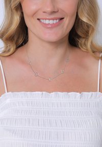 Sølv hjertehalskæde med fem hjerteformede led på en tynd kæde, båret over en hvid, tekstureret off-the-shoulder top.