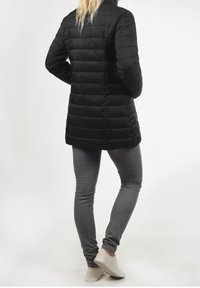 Černý prošívaný kabát s vysokým límcem a vertikálním prošíváním, doplněný šedými úzkými džínami a světlými teniskami.