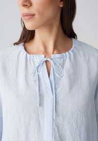 Lichtblauw linnen blouse met een ronde halslijn, strikdetails aan de voorkant en een ontspannen pasvorm. De stof heeft een zachte, gestructureerde uitstraling.