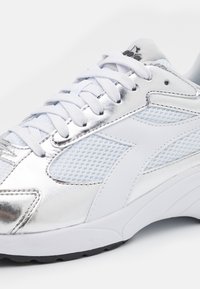 Vit träningsskon med silverfärgad syntetisk ovandel, meshpaneler och vita snören. Har en gummisula och diskret logotyp på tungan.