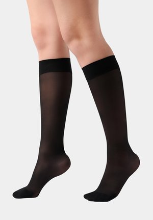 Beine, die durchsichtige schwarze knielange Strümpfe mit festen schwarzen Bündchen oben tragen, gezeigt vor einem schlichten hellen Hintergrund.