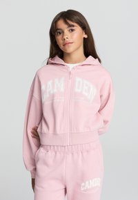 Felpa leggera rosa con zip e cappuccio, caratterizzata da testo stampato bianco. Texture morbida, vestibilità comoda, polsini e orlo a costine, abbinata a pantaloni coordinati.