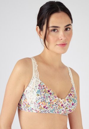 Vrouw met donker haar die een beha met bloemenprint draagt met kanten bandjes en rand, licht naar de zijkant kijkend tegen een effen achtergrond.