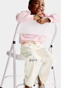 Sudadera rosa clara, pantalones crema con estrellas de mar azules y bordado de "ALOHA", y sandalias plateadas brillantes. Sentado en una silla blanca.