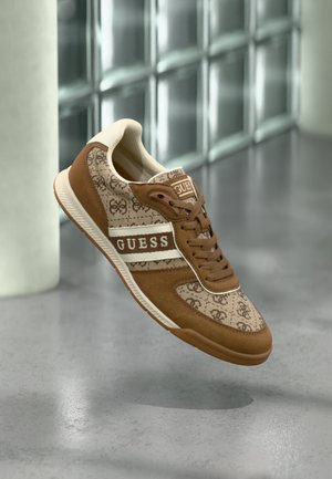 Bruine en beige GUESS sneaker met logoprint en veters, hangend boven een gepolierde betonvloer in een binnenomgeving.