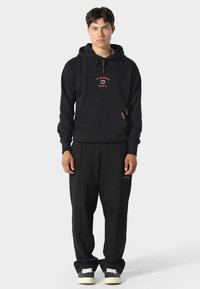 Sudadera negra con bolsillo frontal, con logo rojo bordado. Combinada con pantalones de chándal negros y zapatillas negras con suela blanca.