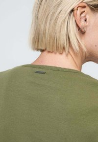 Top en tricot vert olive avec un col rond, présentant un léger jeu de côtes et une petite étiquette métallique à l'arrière. Cheveux courts visibles.