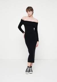 Rochie midi neagră, cu umerii descoperiți, realizată dintr-un material elastic, cu mâneci lungi și un design ajustat, asortată cu adidași negri și albi.