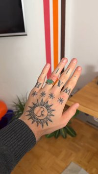 Tetovaná ruka s různými vzory, včetně slunce s paprsky, čar a hvězd. Na prsteníčku je zlatý prsten se zeleným kamenem.