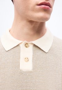 Κοντινό πλάνο ενός ατόμου που φοράει μια μπεζ πλεκτή πόλο μπλούζα με δύο κουμπιά, που φαίνεται το γιακά και μέρος του λαιμού και του πηγουνιού.