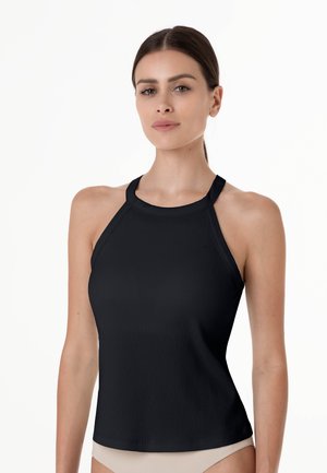 Giovane donna che indossa un top halter nero a coste senza maniche e pantaloni beige, in piedi su uno sfondo bianco semplice.