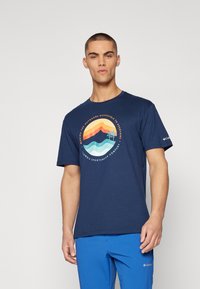 Marineblå kortærmet t-shirt lavet af bomuld, med en flerfarvet grafik af bjerg og bølger samt tekst. Bæret med blå bukser.