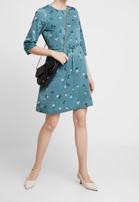 Turkos blommig klänning med trekvartslånga ärmar, snörliv och en utsvängd kjol. Matchad med en svart axelremsväska och beige klackar.