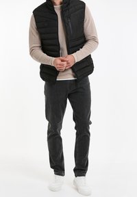 Veste noire gonflée avec col haut, poche à fermeture éclair sur le devant, portée par-dessus un pull côtelé beige clair, associée à un jean foncé et des baskets blanches.