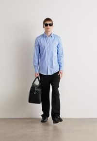 Camisa de botones azul claro, pantalones negros, zapatos negros y gafas de sol. Llevando un bolso de cuero negro con un acento de hardware redondo.
