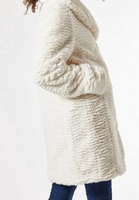 Nepbontjas in crème, met een gestructureerd, geribbeld oppervlak. Het heeft een capuchon en een losse pasvorm, met oversized mouwen.