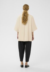 Beige oversized top med korte ærmer, glat tekstur, rygsømsdetalje, parret med sorte tapered bukser og flade sorte sko.