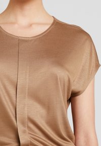 Hnědý, krátký rukáv vršek vyrobený z hladkého materiálu; má kulatý výstřih a jemné faldy na přední části. Minimalistický design.