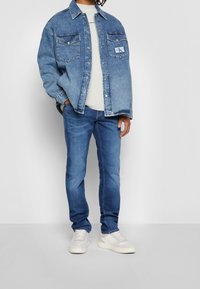 Jeansjacke mit spitzem Kragen und zwei Brusttaschen, über einem hellgrauen Pullover getragen, kombiniert mit blauen Jeans und weißen Sneakern.