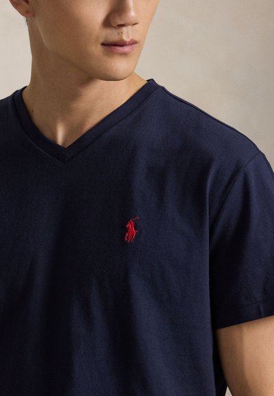 Tamnoplava majica od pamuka s V-izrezom i kratkim rukavima, s malim crvenim izvezenim logotipom na lijevoj strani prsa. Meka tekstura, ležeran dizajn.