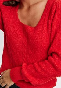 Rot gestrickter Pullover mit tiefem V-Ausschnitt, der strukturierte Zopfmuster und puffige Ärmel aufweist. Glattes, warmes Material und gerippte Bündchen.
