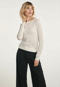 Biała bluzka z długim rękawem, wykonana z lekkiego materiału, o dopasowanym kroju. Posiada ściągacze przy mankietach oraz subtelny akcent z logo na dolnym wykończeniu. Czarne spodnie z szerokimi nogawkami.