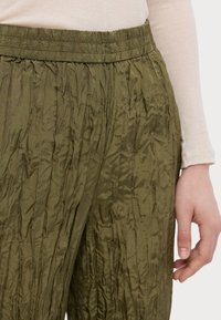 Close-up van een persoon in olijfgroene gekreukt broek met een elastische tailleband en een beige lange mouw top, hand ontspannen aan de zijkant.