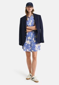 Blå blomstret kjole med korte ermer, lagdelt med en marineblå blazer. Hvite sneakers med marineblå detaljer. Caps med logo.