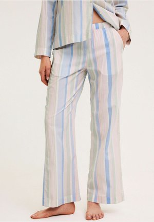 Persoon die losse pyjamabroek met pastelkleurige strepen en bijpassende top draagt, met hand in zak, blootsvoets staand op een effen achtergrond.