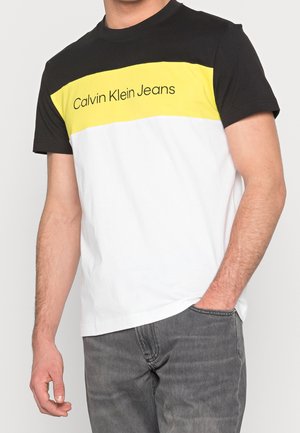 Mężczyzna w koszulce z krótkim rękawem w kolorowe bloki, w kolory czarny, żółty i biały, z napisem "Calvin Klein Jeans", w zestawieniu z szarymi dżinsami.