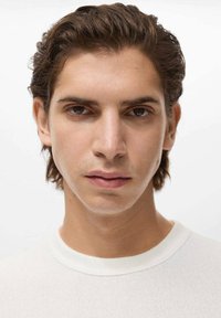 Prim-plan al unui model masculin cu păr șaten deschis și o expresie neutră, îmbrăcat într-un pulover alb, texturat, ajustat. Iluminarea moale se concentrează pe trăsăturile feței.