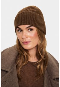 Femeie cu părul castaniu deschis, purtând o căciulă din lână gri cu model, un top maro și un palton din lână maro, privind ușor lateral.