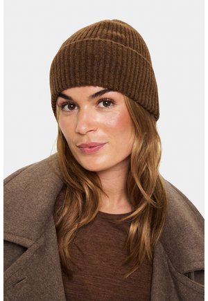 Sieviete ar gaiši brūniem matiem, kas valkā brūnu ribotu adīto cepuri, brūnu krekla augšu un brūnu vilnas mēteli, kas skatās nedaudz uz sāniem.