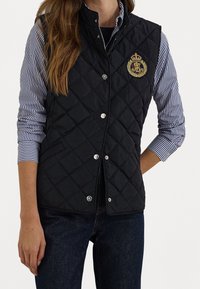 Svart quiltad väst i nylon med silverfärgade tryckknappar, med en guldig broderad logotyp och sidofickor. Bärs över en randig skjorta.