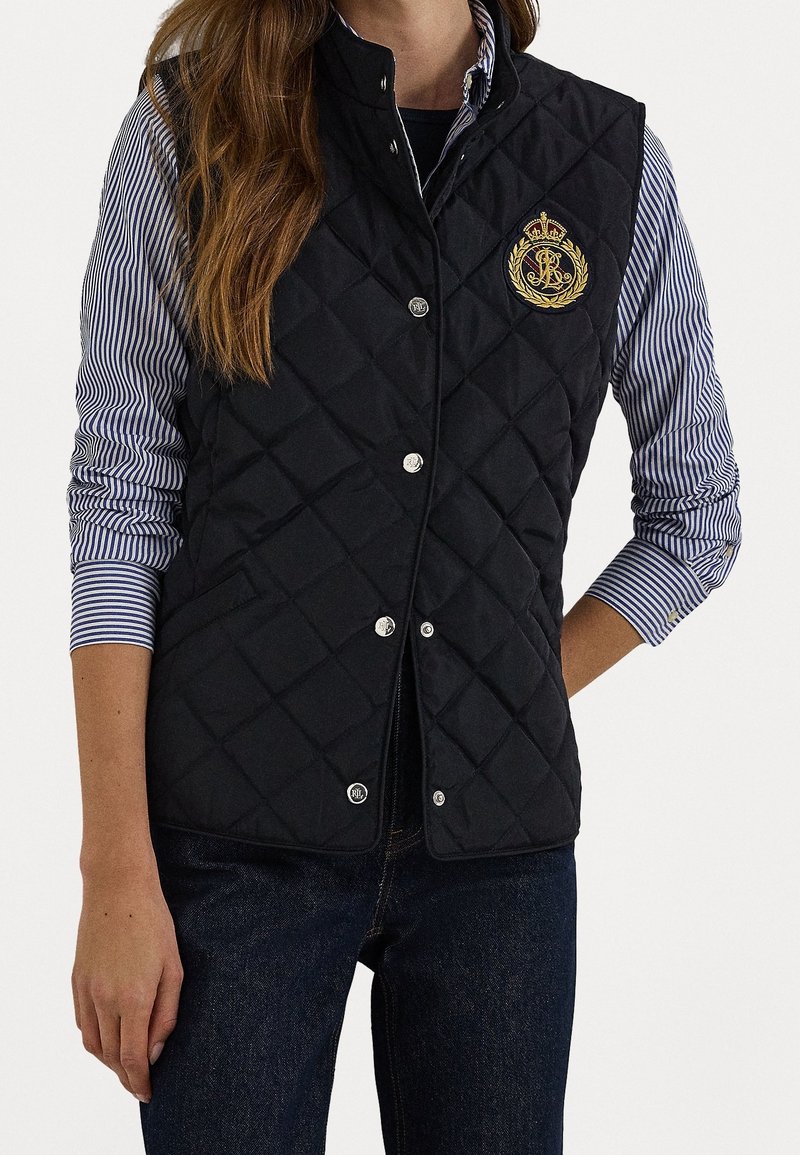 Svart quiltad väst i nylon med silverfärgade tryckknappar, med en guldig broderad logotyp och sidofickor. Bärs över en randig skjorta.
