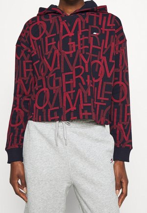 Osoba ubrana w granatową bluzę z kapturem z czerwonym nadrukiem w całości oraz jasnoszare spodnie dresowe ze ściągaczami, na jednolitym tle.