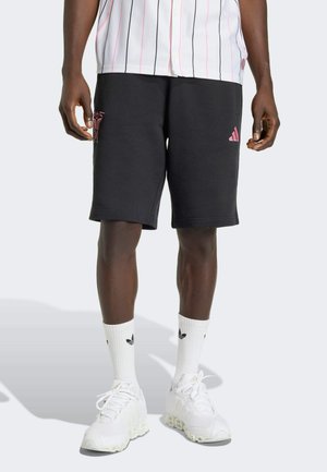 Male wearing black Adidas shorts with pink logos, white high socks, white sneakers, and a white pinstriped shirt standing on a plain background.