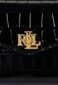 Czarna torebka z lakierowanej skóry ma teksturę przypominającą krokodylą skórę. Na froncie widoczne jest złote zapięcie z logo.
