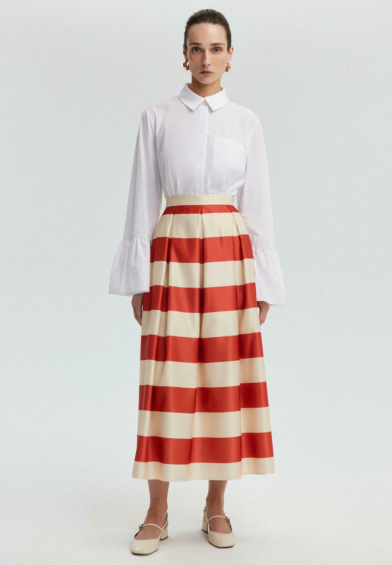 White collared shirt with bell sleeves paired with a long, pleated skirt featuring bold red and cream horizontal stripes. Light shoes complete the look.