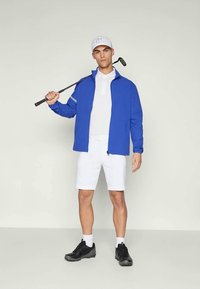 Mann iført blå jakke, hvit polo og shorts, svarte joggesko og hvit caps, holder golfklubbe over skulderen mot en ensfarget bakgrunn.