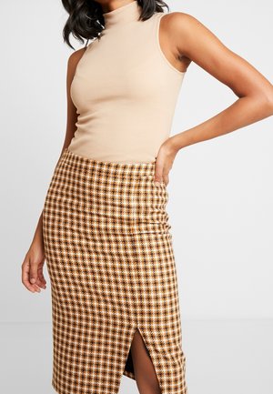 Kobieta w beżowym, bezrękawnym golfie i brązowej spódnicy ołówkowej w kratę z rozcięciem z przodu, pozująca z jedną ręką na biodrze.