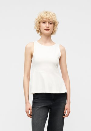 Persona giovane con capelli biondi ricci che indossa un top bianco senza maniche con un peplo e jeans grigio scuro, in piedi davanti a uno sfondo uniforme.