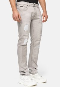 Jeans grigi chiari con toppe strappate, design slim-fit, cinque tasche e dettagli di sbiadimento delicati. Abbinati a sneakers bianche.