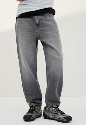 Person trägt lockere graue Jeans und schwarz-graue Sportschuhe und steht vor einem weißen Hintergrund.