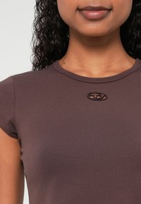 Hnedé priliehavé tričko s krátkymi rukávmi, vyrobené z jemne štruktúrovanej látky a s malým emblémom pri výstrihu.