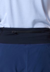 Primo piano della vita di una persona che indossa pantaloni sportivi blu con una tasca nera con cerniera e una maglietta bianca strutturata.