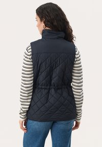 Marineblauer gesteppter Weste mit einem hohen Kragen, tailliert und mit glattem Rücken. Getragen über einem langärmeligen, weiß-blauen gestreiften Hemd.