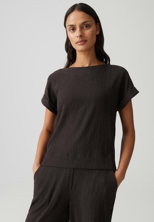 Femme aux longs cheveux foncés portant un haut texturé à manches courtes de couleur sombre et un pantalon assorti, debout les mains dans les poches devant un fond uni.