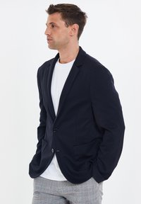 Blazer navy con dos bolsillos frontales, cierre de un botón y solapas con muesca, llevado sobre una camiseta blanca y pantalones con estampado.