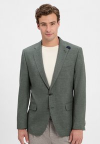 Junger Mann in einem hellgrünen Blazer über einem elfenbeinfarbenen Hemd, mit lockigem Haar und einem leichten Lächeln, posiert vor einem schlichten weißen Hintergrund.